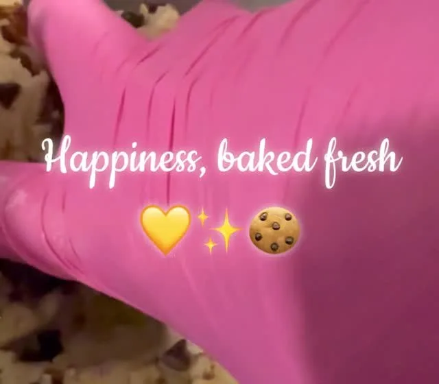 Pink-gloved hand folding chocolate chunk cookie dough in a steel mixing bowl