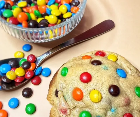 The Happy Chapter cookie studded with colorful M&M’s next to a bowl and spoonful of chocolate candies